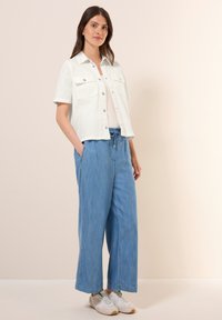 Giacca di jeans bianca, leggera e a maniche corte, con tasche a patta, abbinata a pantaloni blu in chambray larghi e sneaker bianche.