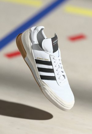 White Adidas Mundial Goal sneaker with black stripes and gum sole, shown mid-air over a sports court floor with colored lines.