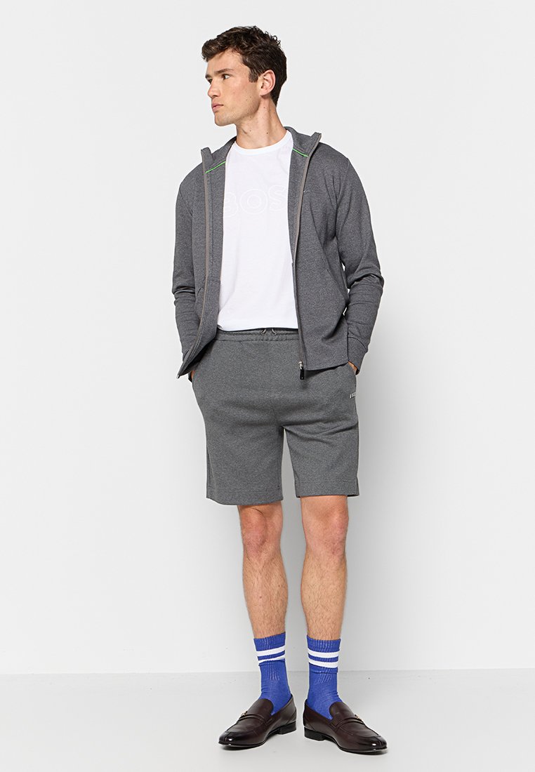 Grijze zip-up hoodie, witte t-shirt, grijze shorts, blauw gestreepte sokken, bruine schoenen. Hoodie heeft groene accenten en voorzakken.