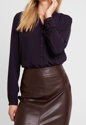 Femme portant un chemisier bleu marine foncé à boutons avec des pois rouges et une jupe en cuir marron taille haute, main près du visage, fond clair.