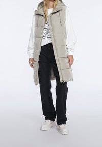 Hellgraue gesteppte Weste mit Kapuze, vertikaler Steppung und Frontreißverschluss, über einem weißen Sweatshirt und schwarzen Hosen mit Cargotaschen.