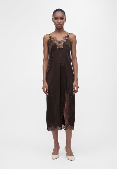 Femme en robe nuisette marron foncé bordée de dentelle avec fines bretelles et talons blancs pointus, debout devant un fond blanc uni.