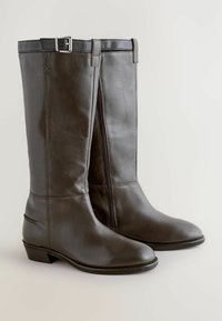 Stivali al ginocchio in pelle marrone scuro con chiusura a zip, tacco basso e una fascia decorativa nella parte superiore. Texture liscia e design aderente.