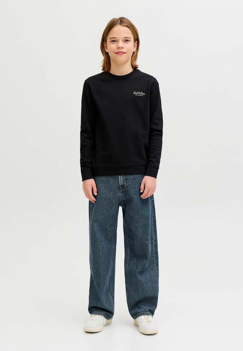 Vállig érő hajú gyermek áll előre nézve, fekete pulóvert, laza kék farmert és fehér sportcipőt visel, egyszínű h áttér előtt.