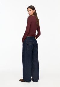 Burgunderfarbene Langarm-Bluse kombiniert mit weit geschnittenen, dunklen Jeans aus Denim mit Gesäßtaschen und einem dezenten, tannfarbenen Lederpatch. Braune Stiefel runden das Outfit ab.