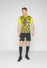 Gul- och svartrandig fotbollsjacka med korta ärmar, tillsammans med svarta shorts som har en logotyp. Svarta strumpor och fotbollsskor fullbordar outfiten.