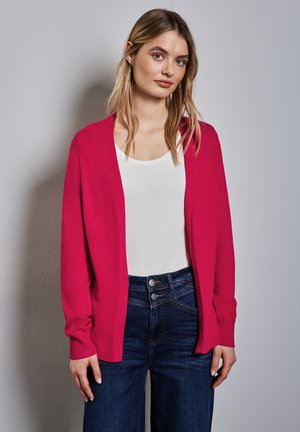 Młoda kobieta o długich blond włosach, nosząca srebrne kolczyki koła, biały top, jasnoróżowy kardigan oraz ciemnoniebieskie dżinsy z wysokim stanem, stoi na tle szarej ściany.