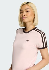 Jasnoróżowa koszulka z brązowym prążkowanym kołnierzem i trzema brązowymi paskami na rękawach. Na piersi znajduje się małe brązowe logo Adidas.
