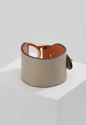Large bracelet en cuir beige avec boucle circulaire dorée et fermeture par bouton, présenté sur un socle blanc avec un fond neutre.