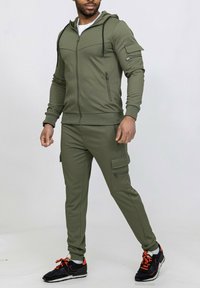 Ensemble de survêtement vert olive avec un sweat à capuche zippé et un pantalon cargo. Comprend des poches et des poignets côtelés. Associé à des chaussures de sport noires et orange.
