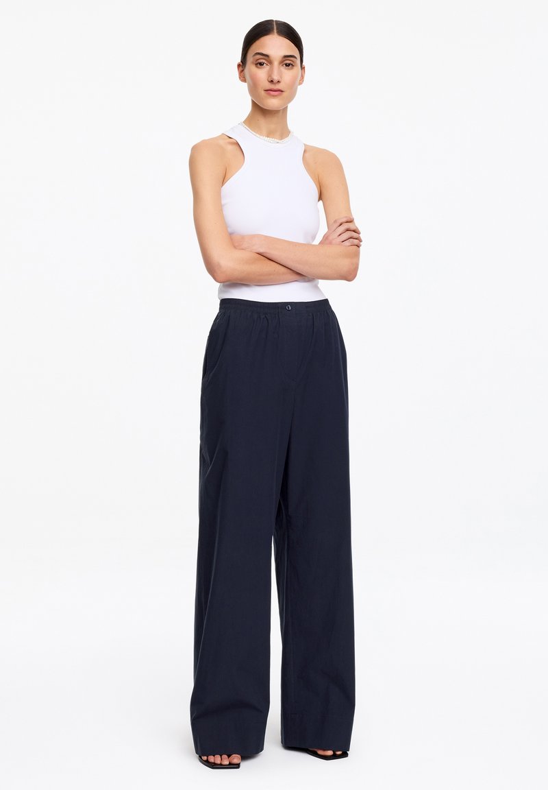 Femme debout, les bras croisés, portant un haut blanc sans manches et un pantalon noir à jambes larges sur un fond blanc uni.