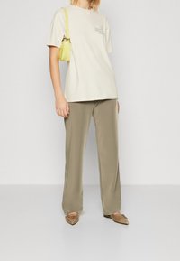 Beige katoenen t-shirt met bedrukte tekst, gecombineerd met olijfgroene, gladde stoffen broek. Model draagt tan puntige flats en houdt een kleine gele tas vast.