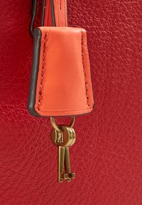 Szczegół czerwonej skórzanej torby z pomarańczową skórzaną metką z czarnymi krawędziami, przymocowanymi kluczami w złotym odcieniu oraz teksturowaną powierzchnią.