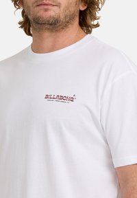 Mann trägt ein weißes Billabong-T-Shirt mit kleinem maroon Logo und dem Text "Feelin' Free Since 73" auf der linken Brust.