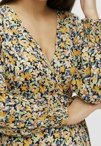 Sukienka w kwiaty z czarnym tłem i kolorowymi żółtymi, białymi oraz fioletowymi kwiatami; dekolt w kształcie v, obszerne rękawy i gumiaki przy mankietach.