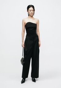 Haut noir sans bretelles associé à un pantalon noir à jambes larges. Le mannequin tient un petit sac à main noir. Les chaussures ont des bouts pointus et un design élégant.