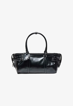 Sac à main en cuir noir brillant avec deux poignées et une étiquette décorative en forme de cœur cousue attachée à une poignée.