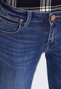 Los jeans de mezclilla azul oscuro presentan un botón de cobre, un diseño de cinco bolsillos y un ligero desgaste en la parte frontal. Camisa a cuadros visible arriba.
