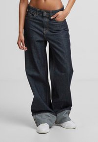 Plačios juodos džinsinės kelnės su oranžinėmis siūlėmis. Yra aukštas liemuo, gilių kišenių ir apvynioti mėgintuvėliai. Derinamos su baltais sportbačiais.