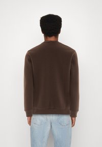 Sudadera marrón con cuello redondo, mangas largas y puños acanalados. Textura lisa, corte relajado, combinada con jeans de un azul claro.