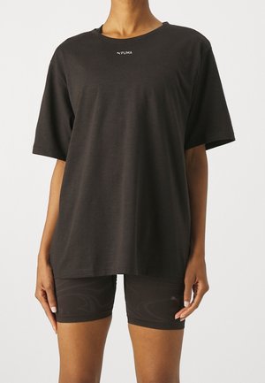 Person trägt ein lockeres schwarzes Puma-T-Shirt und enganliegende schwarze Shorts und steht vor einem schlichten hellen Hintergrund.