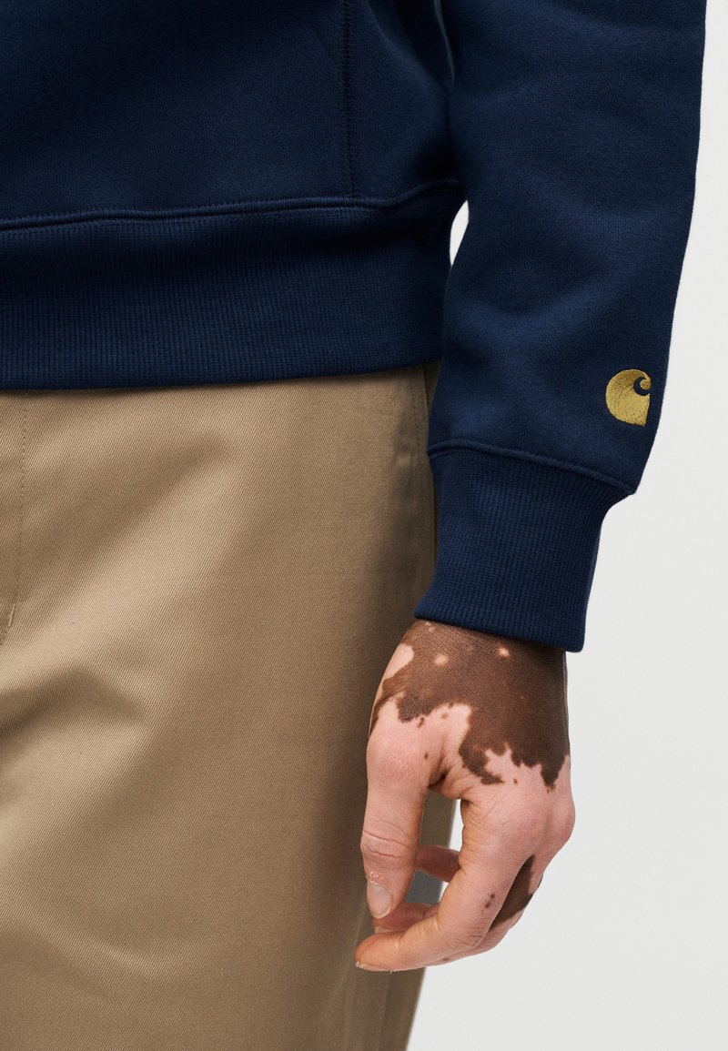 Sweat-shirt bleu marine à poignets côtelés, avec un logo doré sur la manche gauche. Porté avec un pantalon beige, montrant une main avec un motif de peau distinct.