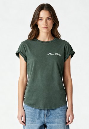 Femme aux longs cheveux bruns portant un t-shirt vert foncé à manches retroussées avec le texte « Miss Paris » et un jean bleu clair, debout sur fond blanc.