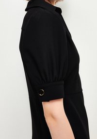 Vestido negro con un escote con cuello, mangas abullonadas y acentos en los puños con herrajes dorados. Textura suave y silueta ajustada.