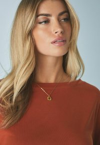 Bronzen top met een ronde halslijn; gladde textuur. Gouden ketting met een vierkante hanger en een schakelketting. Haar in losse golven gestyled.