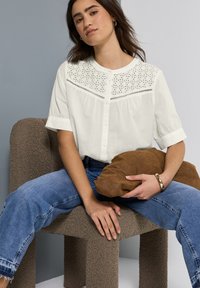 Bílá košile s knoflíky a designem broderie anglaise na živůtku, spárovaná s modrými džíny. Drží hnědý sametový polštář. Texturované pozadí.