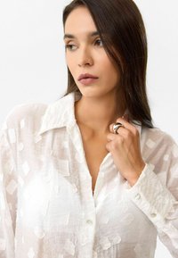 Vrouw met lang bruin haar die een gestructureerd wit overhemd met knopen draagt, de kraag met haar rechterhand vasthoudend en een zilveren ring vertonend.