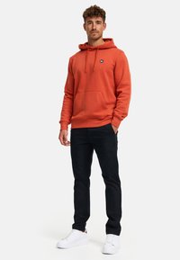 Hoodie arancione ruggine con tasca frontale, cappuccio regolabile con coulisse e logo in evidenza. Abbinato a pantaloni neri e sneakers bianche. Texture liscia.