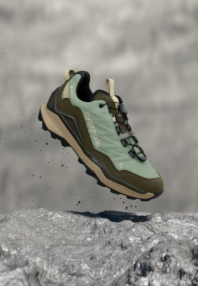 Green and brown hiking shoe with black sole and toggle laces, suspended above rocky ground with dust particles in motion.