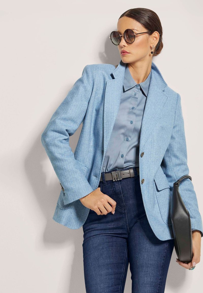 Hellblaue Fischgrät-Blazer über einem blauen Hemd und dunkler Jeans. Accessoires mit schwarzem Gürtel und Handtasche, dazu runde Sonnenbrille.