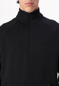 Veste zippée noire avec un col montant, fabriquée en tissu lisse. Présente un logo brodé discret sur la poitrine et des manches raglan.