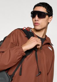 Καφέ ελαφρύ μπουφάν με φερμουάρ και ελαστικές μανσέτες, συνδυασμένο με μεγάλα μαύρα γυαλιά ηλίου. Ορατά λουριά σακιδίου στον ώμο.