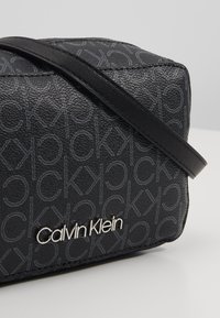 Černá taška z texturovaného syntetického materiálu s potištěným vzorem loga a stříbrným emblémem Calvin Klein, s tenkým černým ramenním popruhem.
