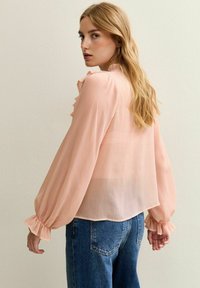 Blusa transparente de color durazno con acentos de volantes en los hombros y puños, mangas largas y un dobladillo redondeado. Combinada con jeans azules.