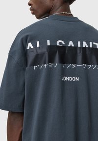Tamsiai pilkas didelio dydžio marškinėlis su juodu horizontaliu dryžiu ant nugaros, kurioje yra balti užrašai: "ALLSAINTS" ir "LONDON." Minkšta medvilnės tekstūra.
