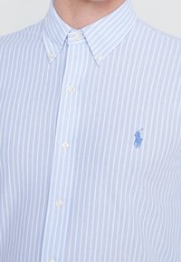 Jasnoniebieska koszula w paski na guziki wykonana z lekkiego materiału, z klasycznym kołnierzem i małym haftowanym logo na lewej piersi.