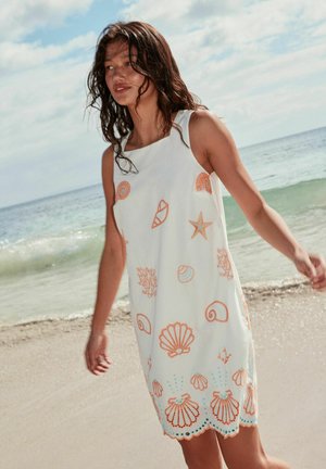 Woman with wavy hair wearing a white sleeveless dress with orange seashell and sea life prints, walking along a sandy beach with ocean waves.