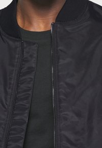 Giacca bomber nera con tessuto liscio e lucido, colletto a coste e chiusura con cerniera. Indossata sopra una maglietta verde scuro.