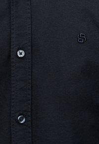 Κοντινό πλάνο ενός σκούρου μπλε πουκαμίσου με δύο ορατά κουμπιά και ένα μικρό κεντημένο λογότυπο στο στήθος.