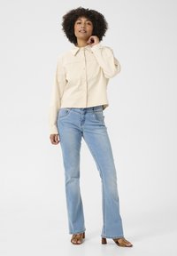 Ljusbeige kortärmad skjorta med knappar och långa ärmar, kombinerad med ljusblå utsvängda jeans och bruna klacksandaler. Vit bakgrund.