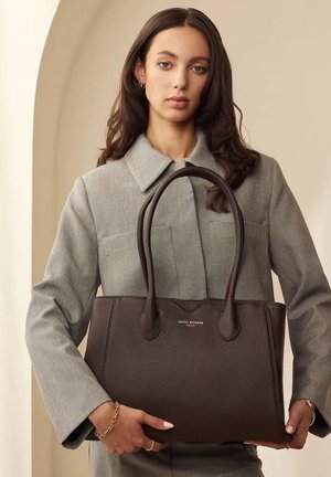 Brown textured leather tote bag with dual handles and simple branding on the front. Model holds it against a neutral background.