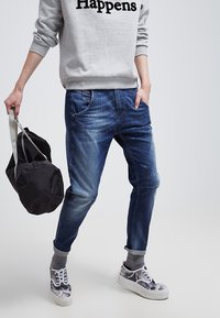 Vapaa-ajan asu, johon kuuluu tummansiniset farkut, joissa on vaalennuksia, harmaa collegepaita mustalla tekstillä sekä kuvioidut harmaat lenkkarit. Mustaa matkakassia pidetään kädessä.