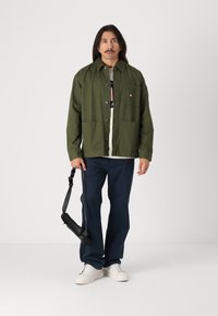Veste en coton vert avec fermeture à boutons, deux poches avant, portée sur un t-shirt blanc. Un pantalon bleu marine et des baskets blanches complètent la tenue.