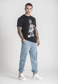 Černé grafické tričko s potiskem karikatury králíka, kombinované se světle modrými cargo kalhotami a bílými teniskami s texturovaným povrchem.