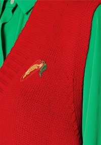 Veste rouge en tricot avec texture côtelée, ornée d'un piment brodé jaune et vert sur le côté gauche, portée sur une chemise verte.