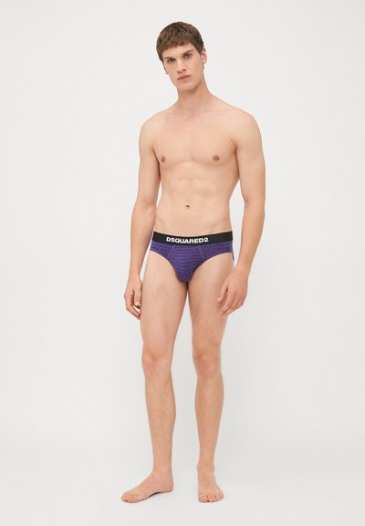 Fialové spodné prádlo s čiernym pásom s logom "DSQUARED2". Priľnavý dizajn s hladkou textúrou. Model stojí bosý na jednofarebnom pozadí.